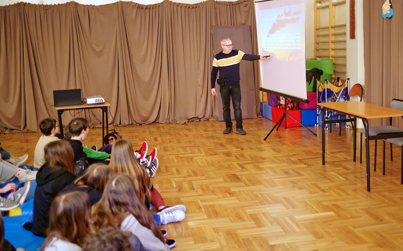 Pracownik „RADPEC” S.A. prowadzi lekcję ciepła dla uczniów klasy VI jednej z radomskich szkół. Stoi przed grupą uczniów. W tle znajduje się ekran z jednym ze slajdów prezentacji.