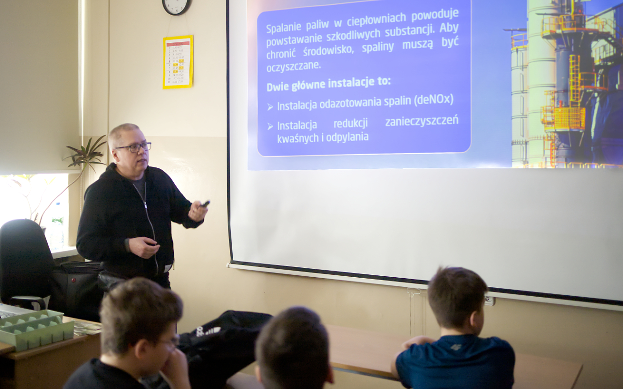 Pracownik „RADPEC” S.A. prowadzi lekcję ciepła dla uczniów klasy VI jednej z radomskich szkół. Stoi przed grupą uczniów. W tle znajduje się ekran z jednym ze slajdów prezentacji.