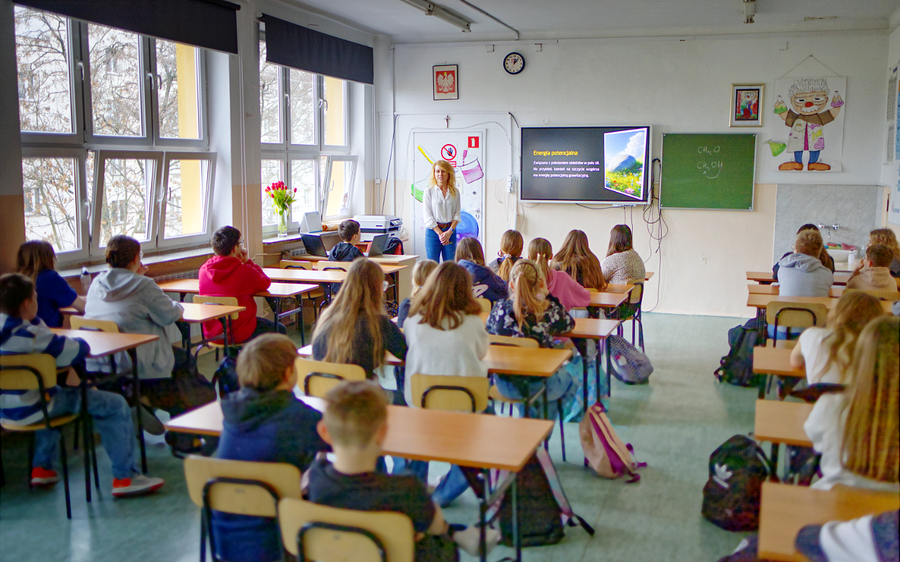 Pracownik „RADPEC” S.A. prowadzi lekcję ciepła dla uczniów klasy VI jednej z radomskich szkół. Stoi przed grupą uczniów. W tle znajduje się ekran z jednym ze slajdów prezentacji.