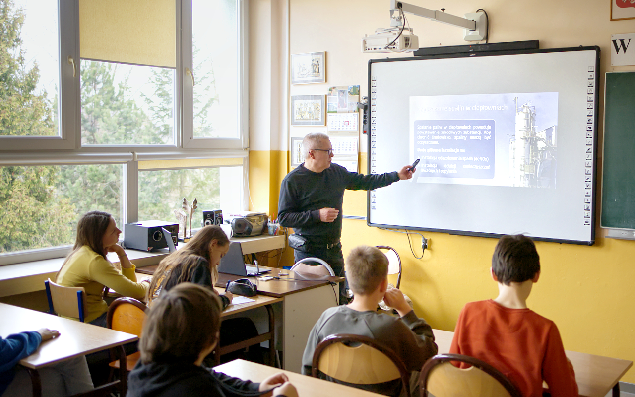 Pracownik „RADPEC” S.A. prowadzi lekcję ciepła dla uczniów klasy VI jednej z radomskich szkół. Stoi przed grupą uczniów. W tle znajduje się ekran z jednym ze slajdów prezentacji.